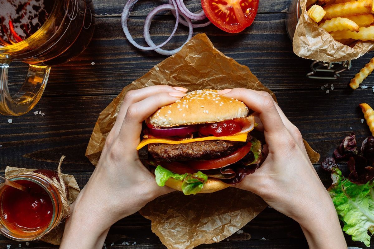 A person is holding a hamburger in their hands on a wooden table.