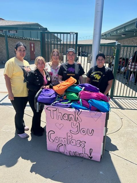 People standing behind a pink thank-you sign with colorful backpacks in front of a fenced entrance.
