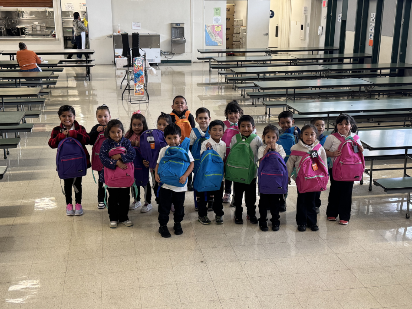 A group of children with colorful backpacks standing in a cafeteria or school lunchroom.