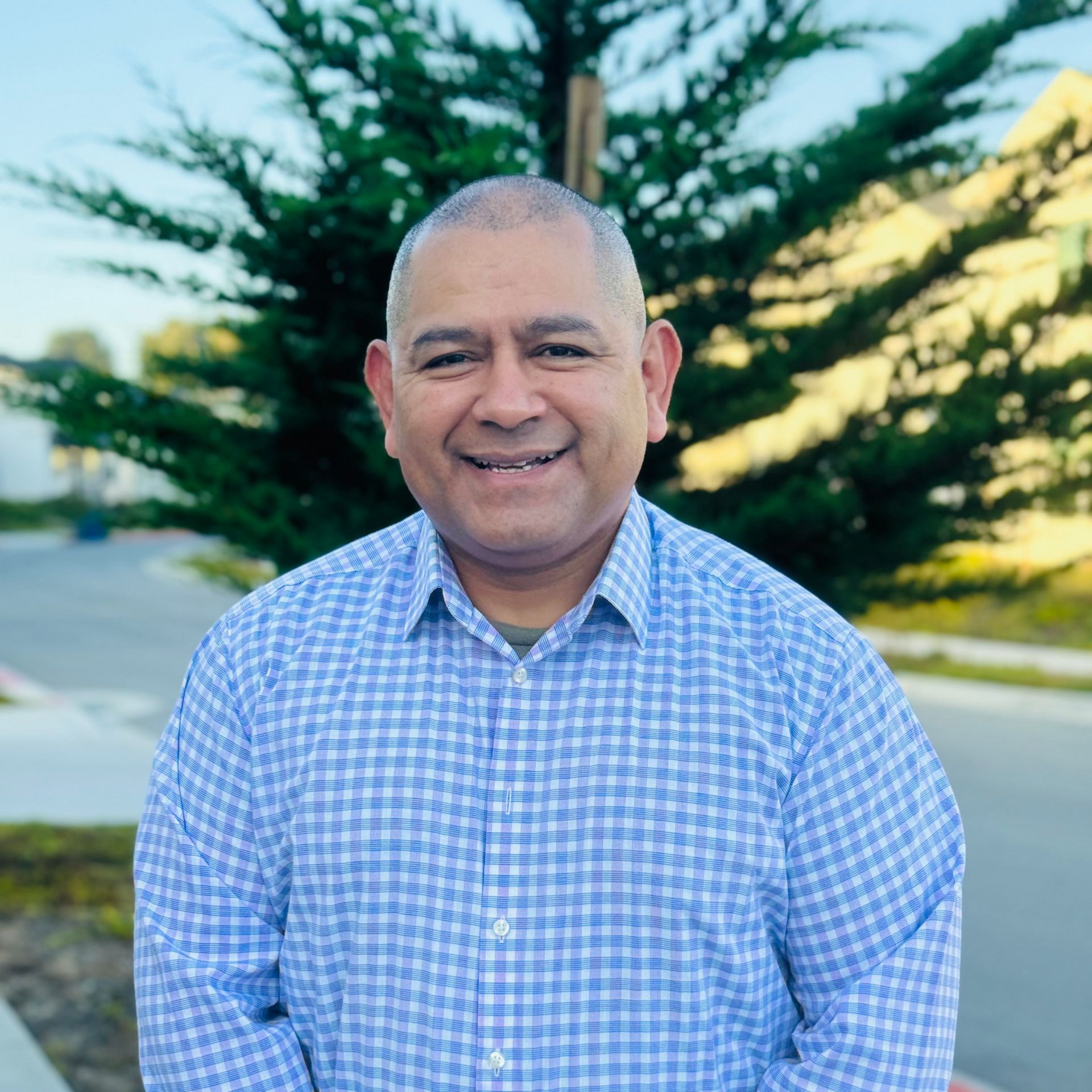 A man wearing a blue and white checkered shirt smiles in front of green shrubbery.