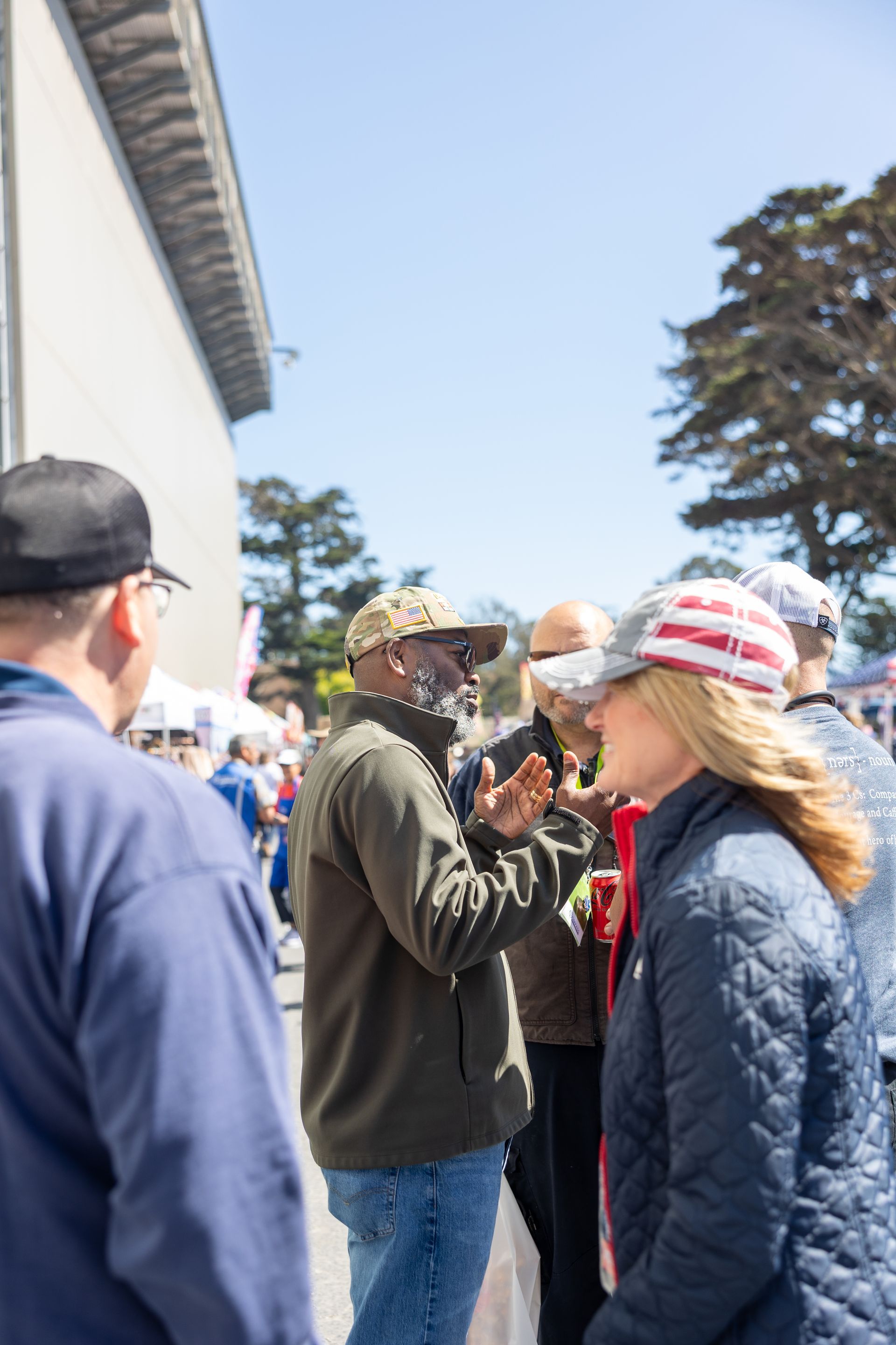People conversing outdoors in front of a building; one man gestures with hands, others wear hats; sunny day.
