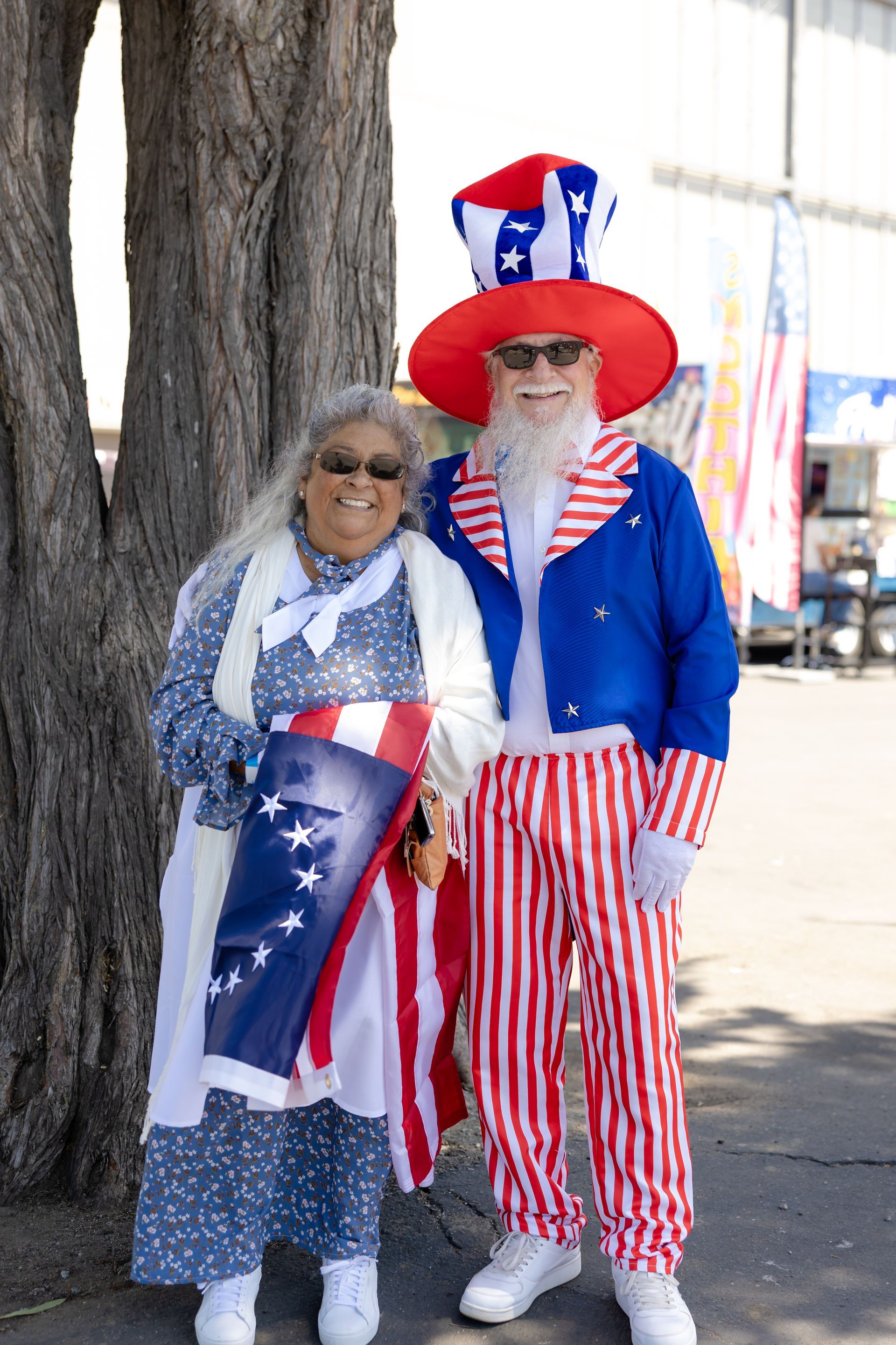 A couple dressed in patriotic costumes, posing outdoors; one as Uncle Sam, the other in a blue dress.