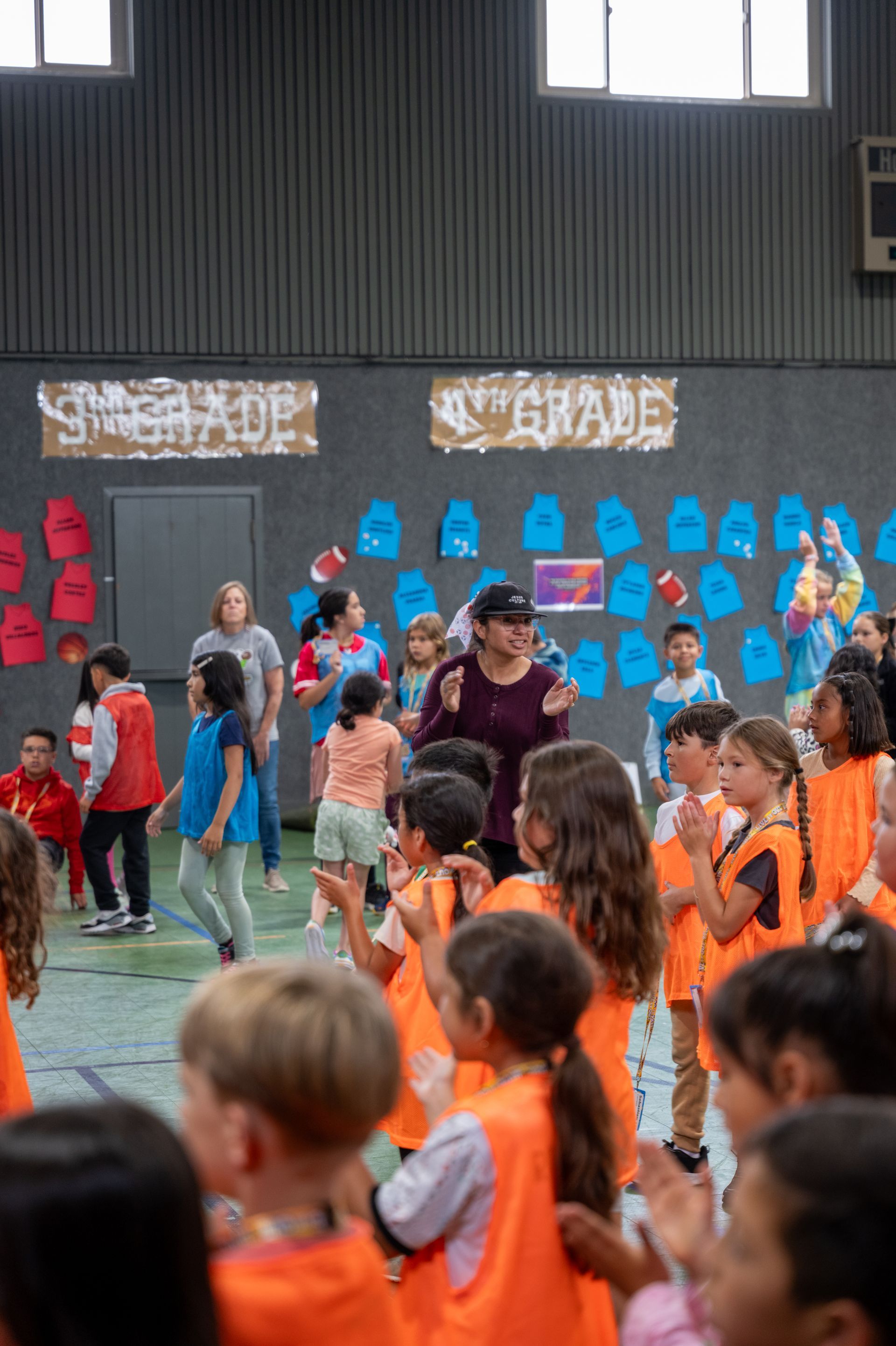 Students in orange vests clap as an adult speaks in a gym.  