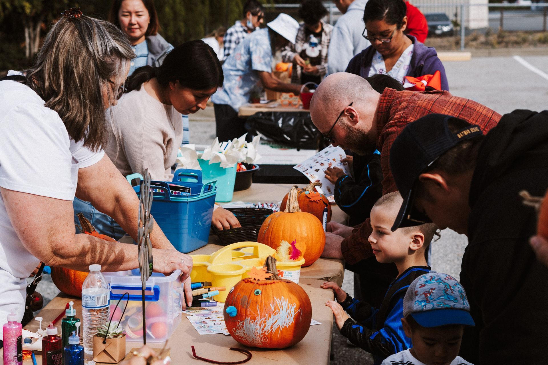 People decorate pumpkins at an outdoor event, some gathered around a table with supplies.