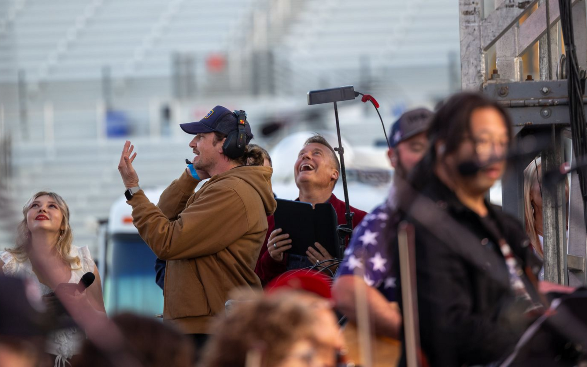 Crew members on a stage: a man in a brown jacket gestures, others smile, looking up.