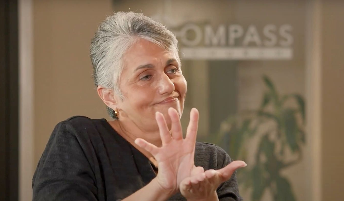 Woman gesturing with hands, seated, smiling. Background features the word