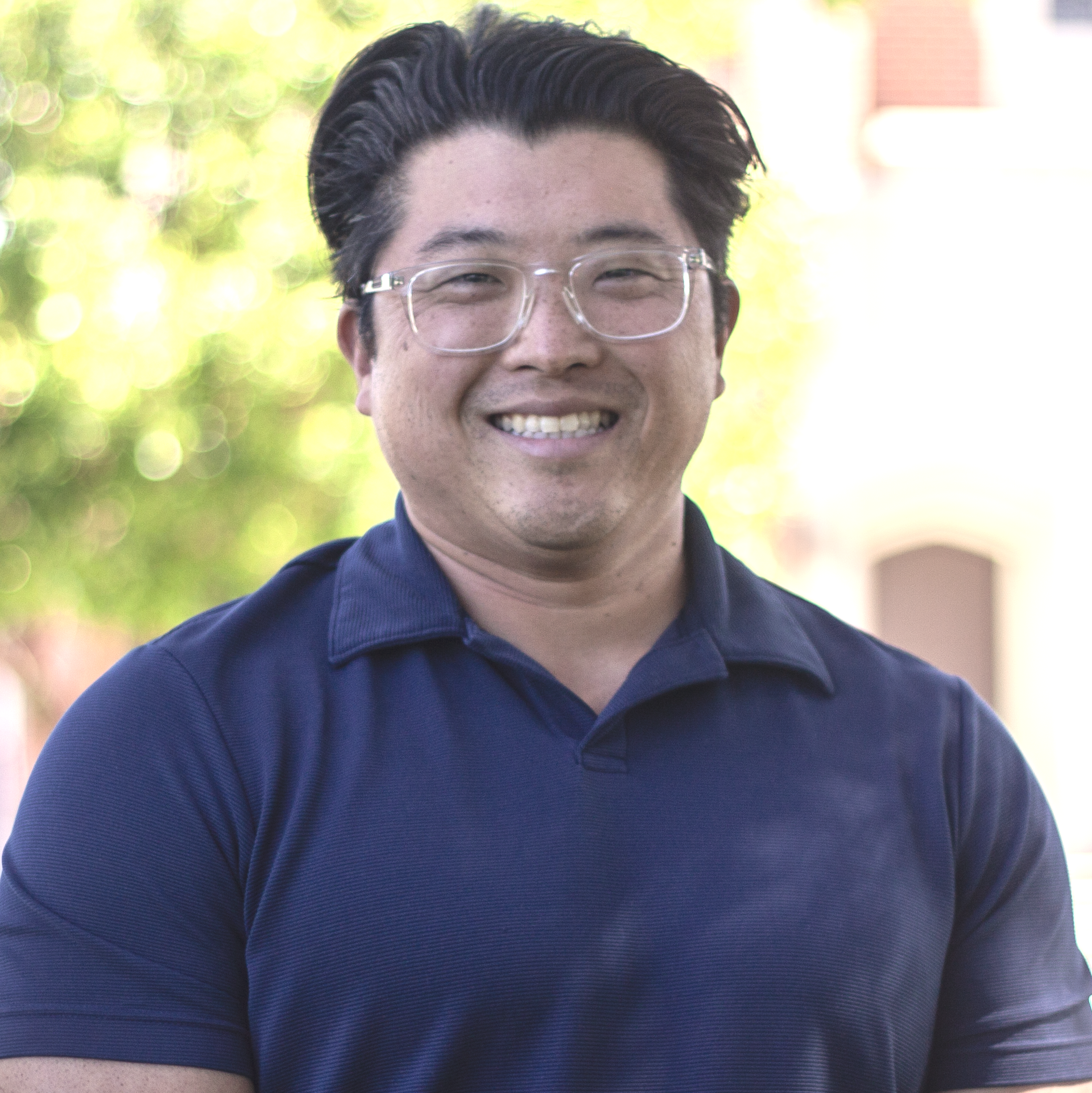 Smiling man wearing glasses and a blue polo shirt outdoors.