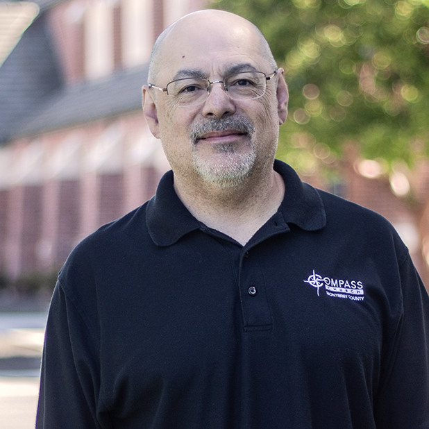 Man with glasses and a goatee in a black polo shirt, with a building in the background.