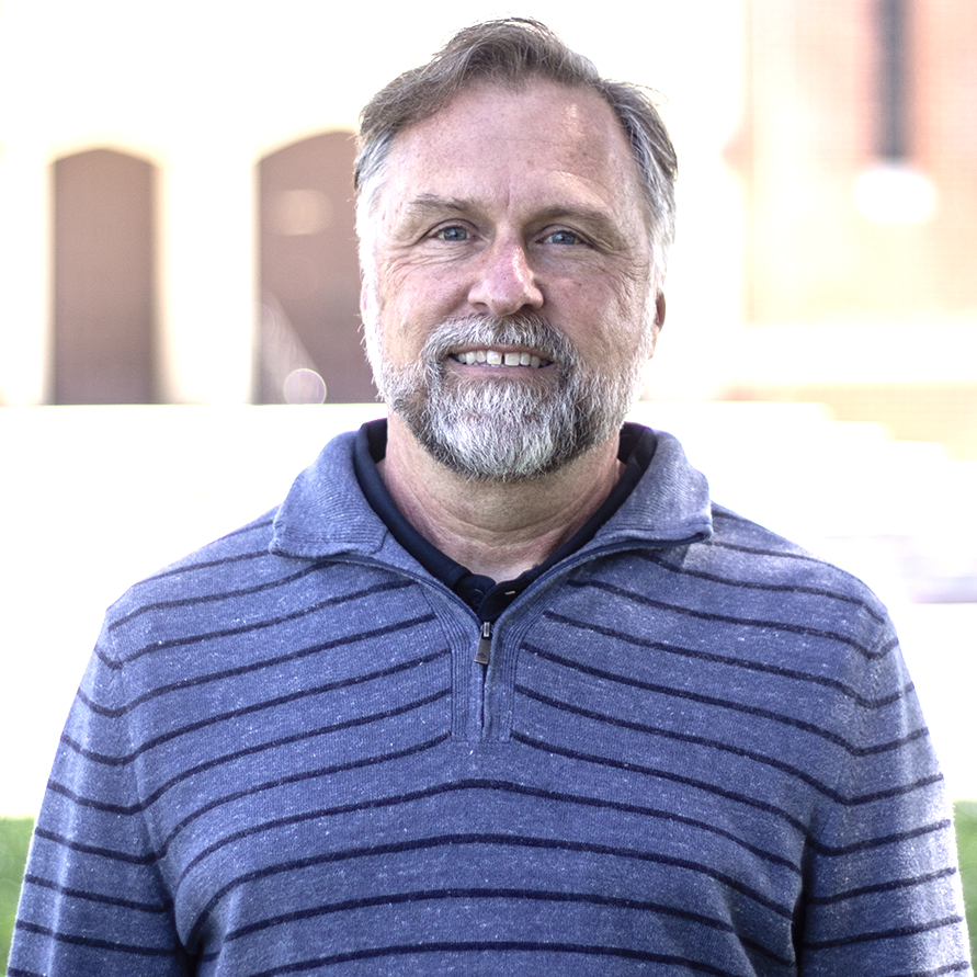 A man with a graying beard and a blue striped sweater smiles outdoors.