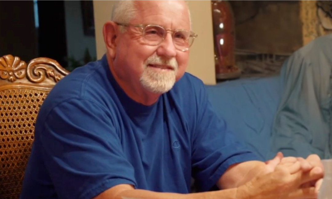 Elderly man with glasses and a white beard smiles, wearing a blue shirt, seated at a table.
