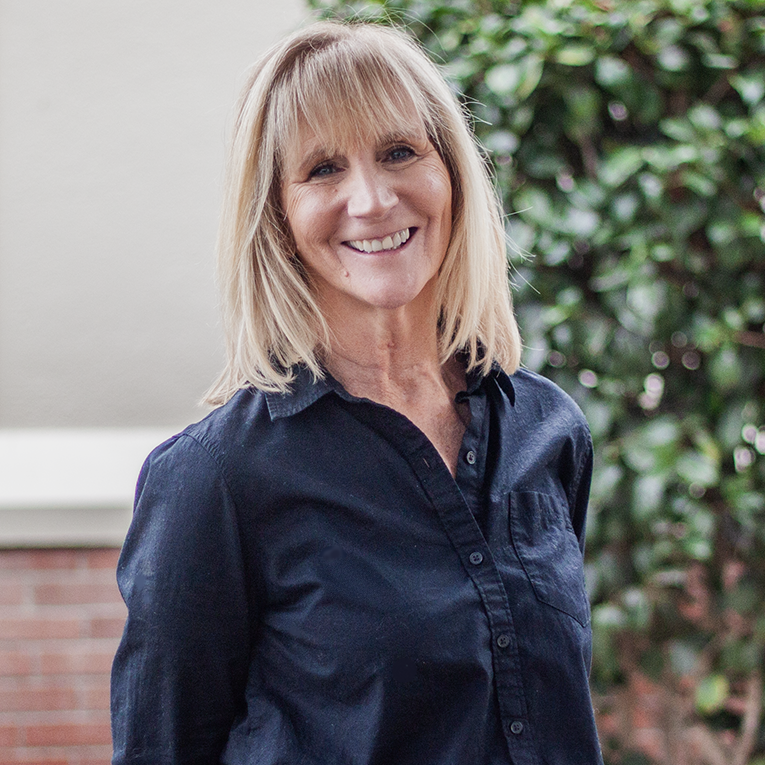 Man with glasses and a graying beard, wearing a blue vest and patterned shirt, smiling outdoors.