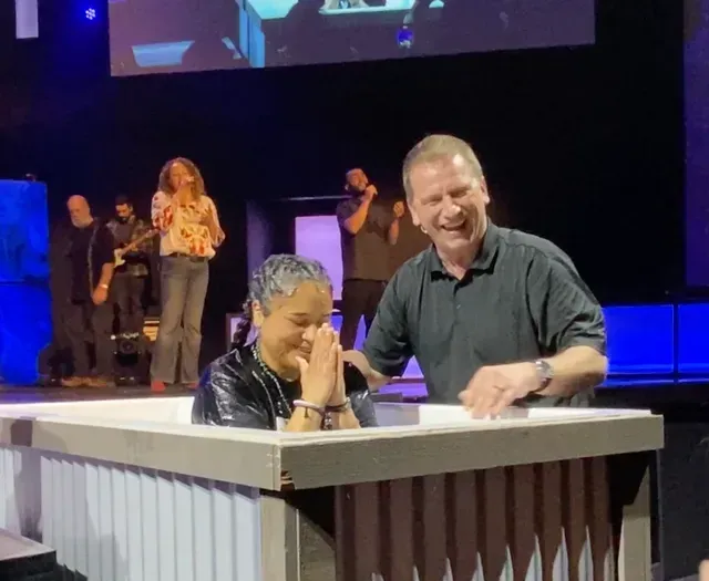 A woman is being baptized by Pastor Todd of Compass Church, Salinas, in a white pool, both smiling on stage with a band.