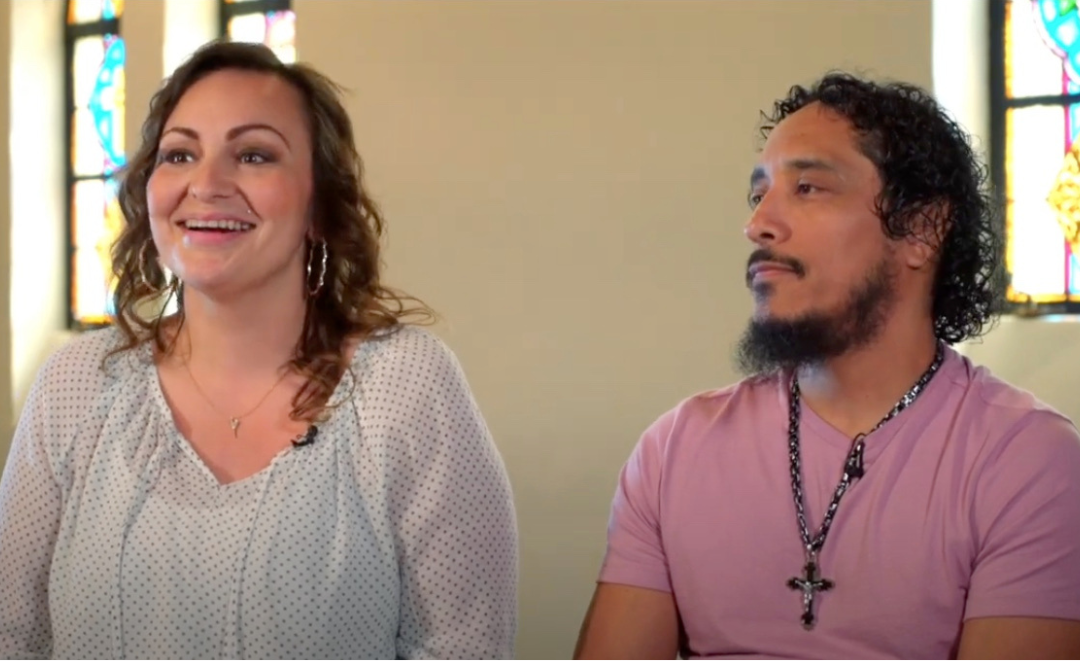 Woman and man smiling, possibly in a church. Man wearing a cross necklace. They appear to be in a video.