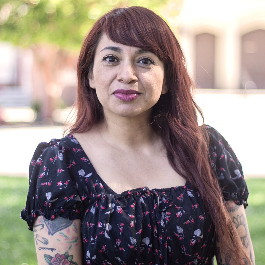 Woman with red hair and tattoos, wearing a floral top, smiling in an outdoor setting.