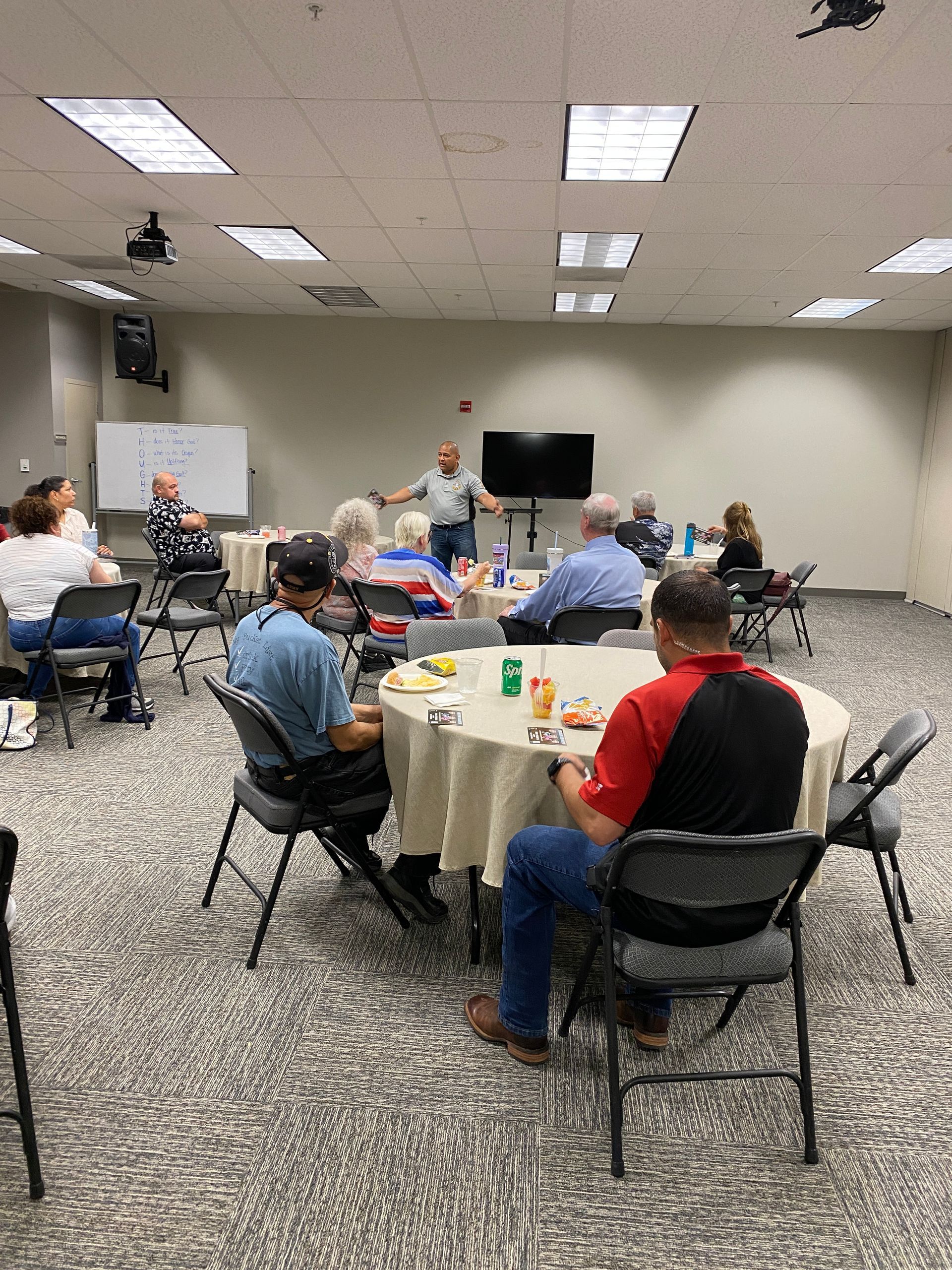 People seated at tables in a room, listening to a man speaking at the front near a TV and whiteboard.