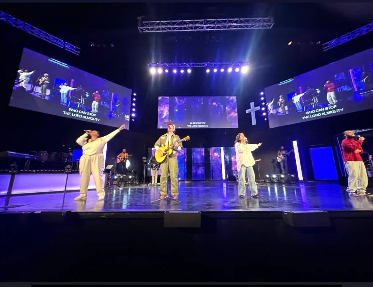 A worship band performs on stage with screens displaying lyrics and visuals.
