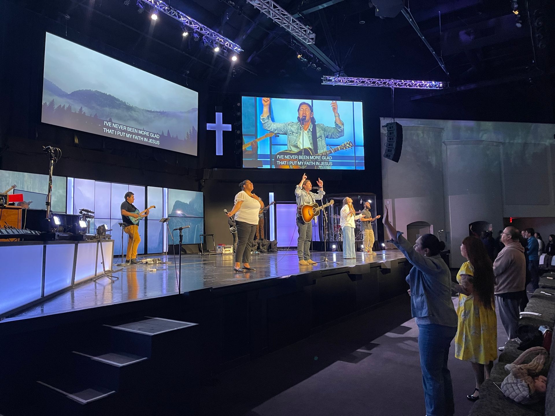 Worship service with band on stage, singing. Audience members stand with hands raised. Large screens display lyrics.