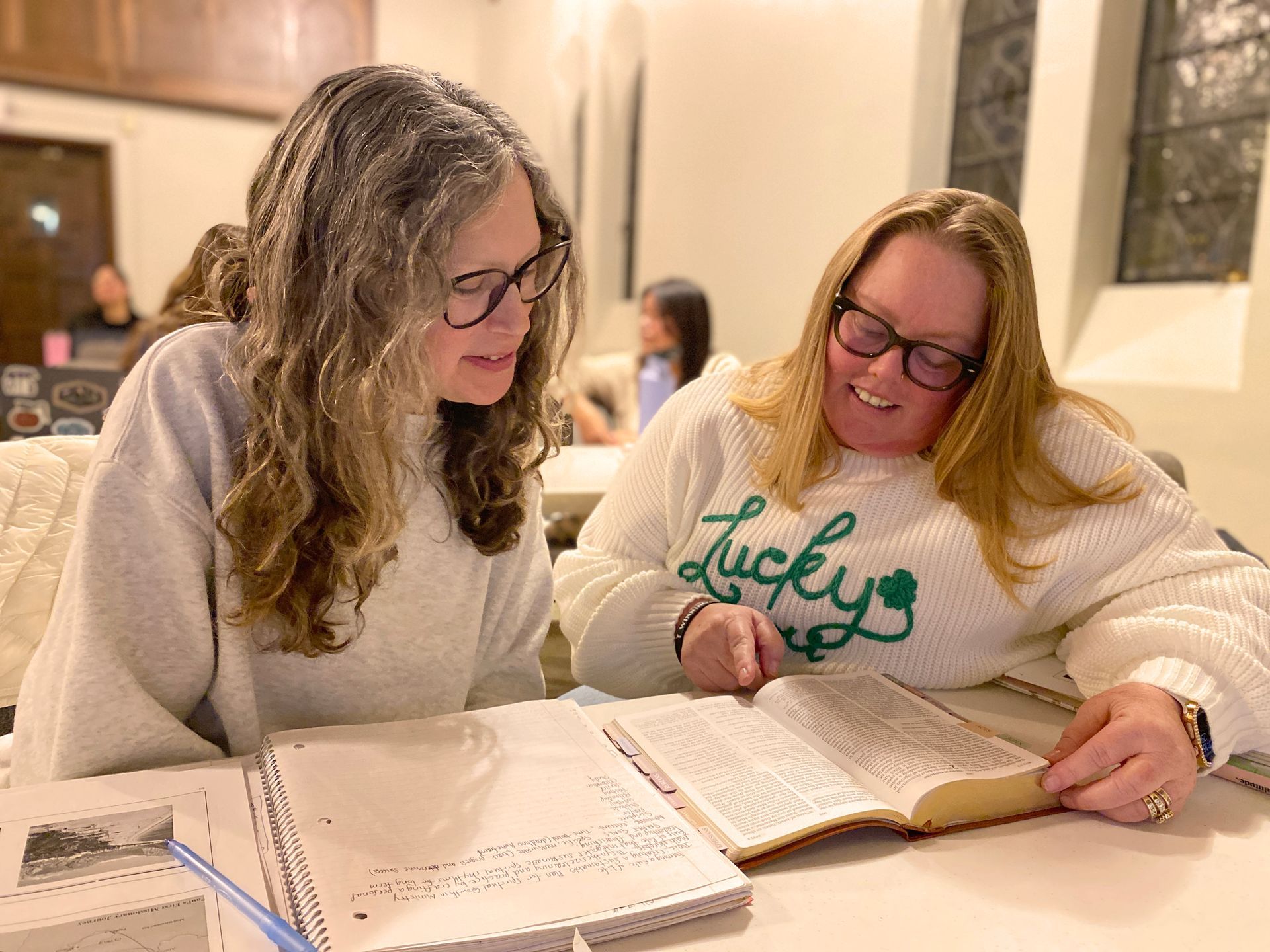 Group of people standing in a room, in front of a raised platform. They are dressed casually, smiling. Tables are in the foreground.