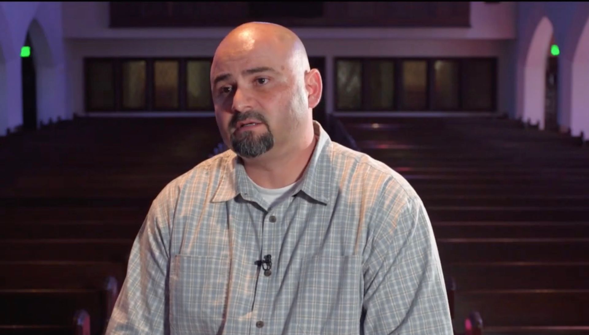 Man in plaid shirt speaking in a church. Wooden pews in background.