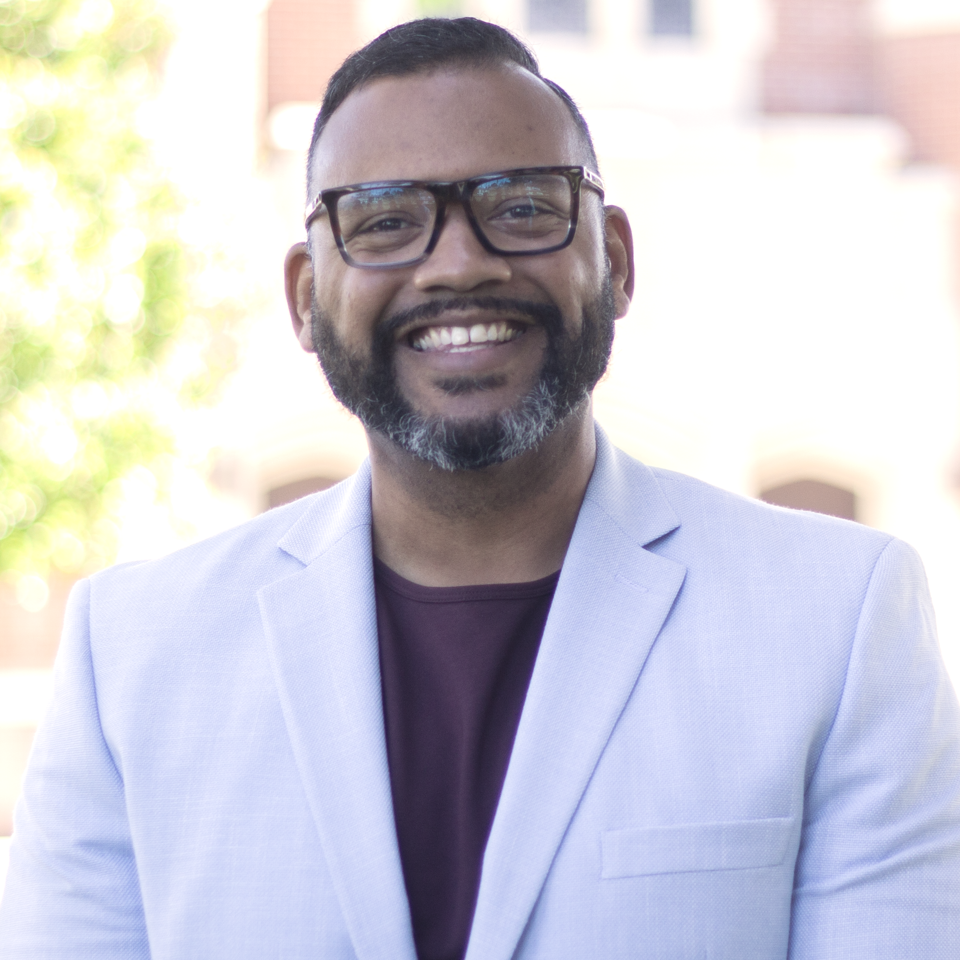 Man in glasses, smiling, wearing a blue jacket over a purple shirt. Building backdrop.