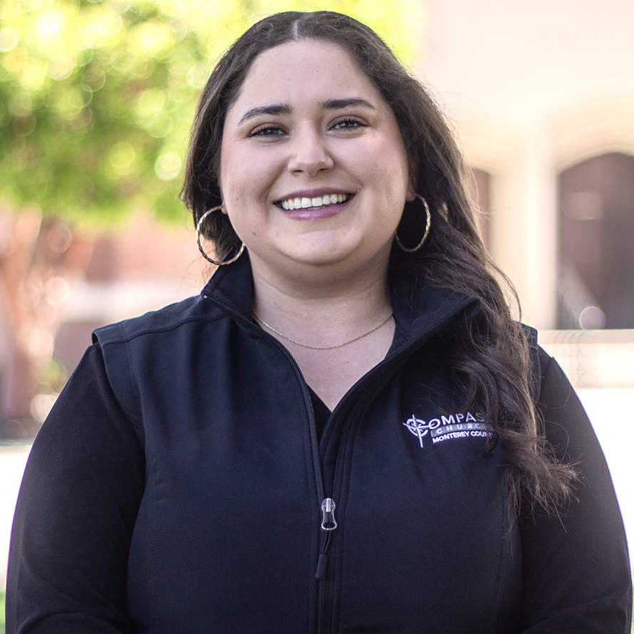 Woman in black vest smiling; outdoor setting with green foliage and building in background.