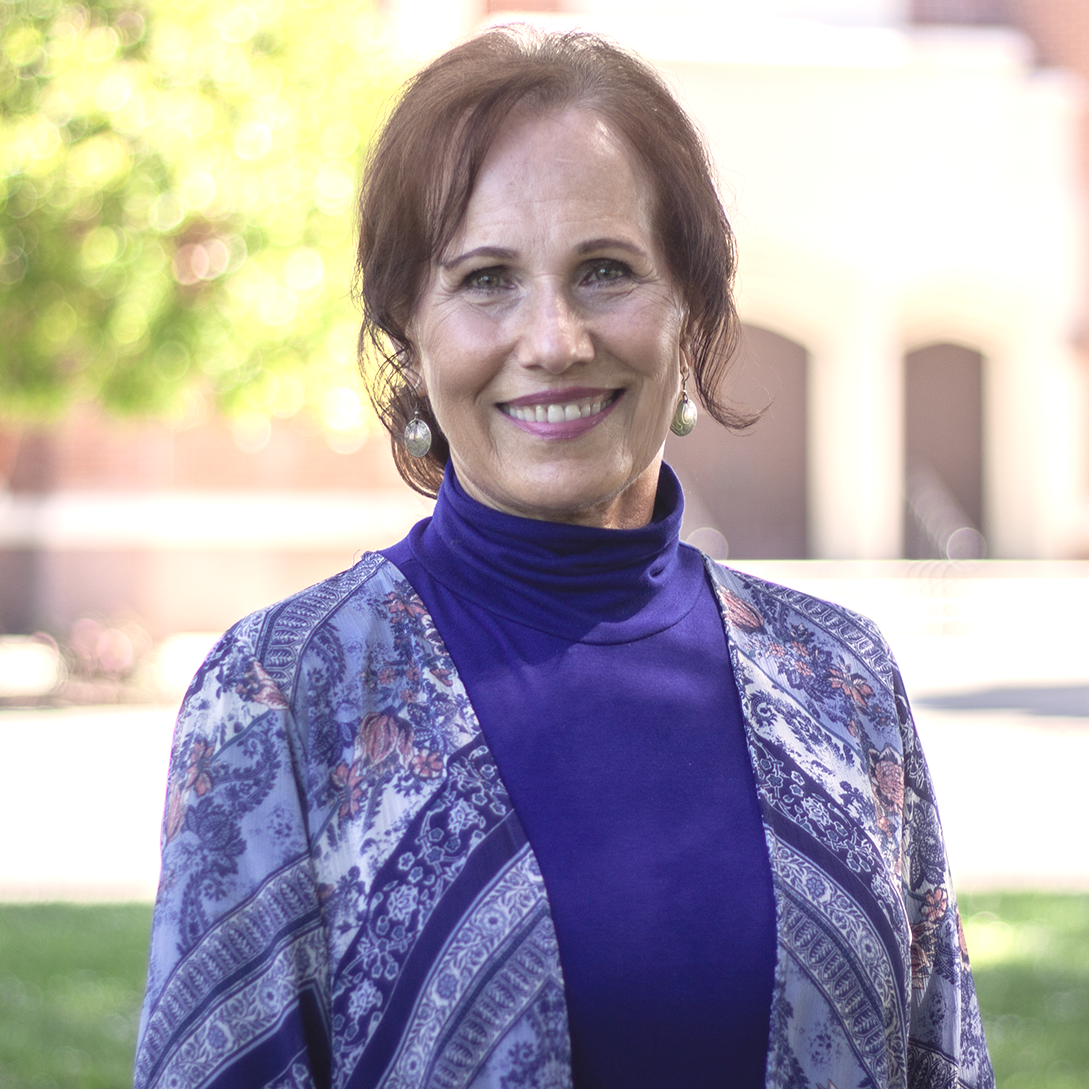 A woman in a blue turtleneck and printed shawl smiles outside near a building.