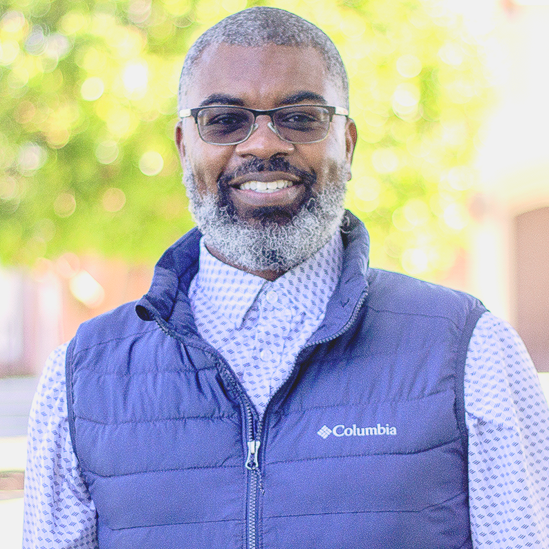 Man with glasses and a graying beard, wearing a blue vest and patterned shirt, smiling outdoors.