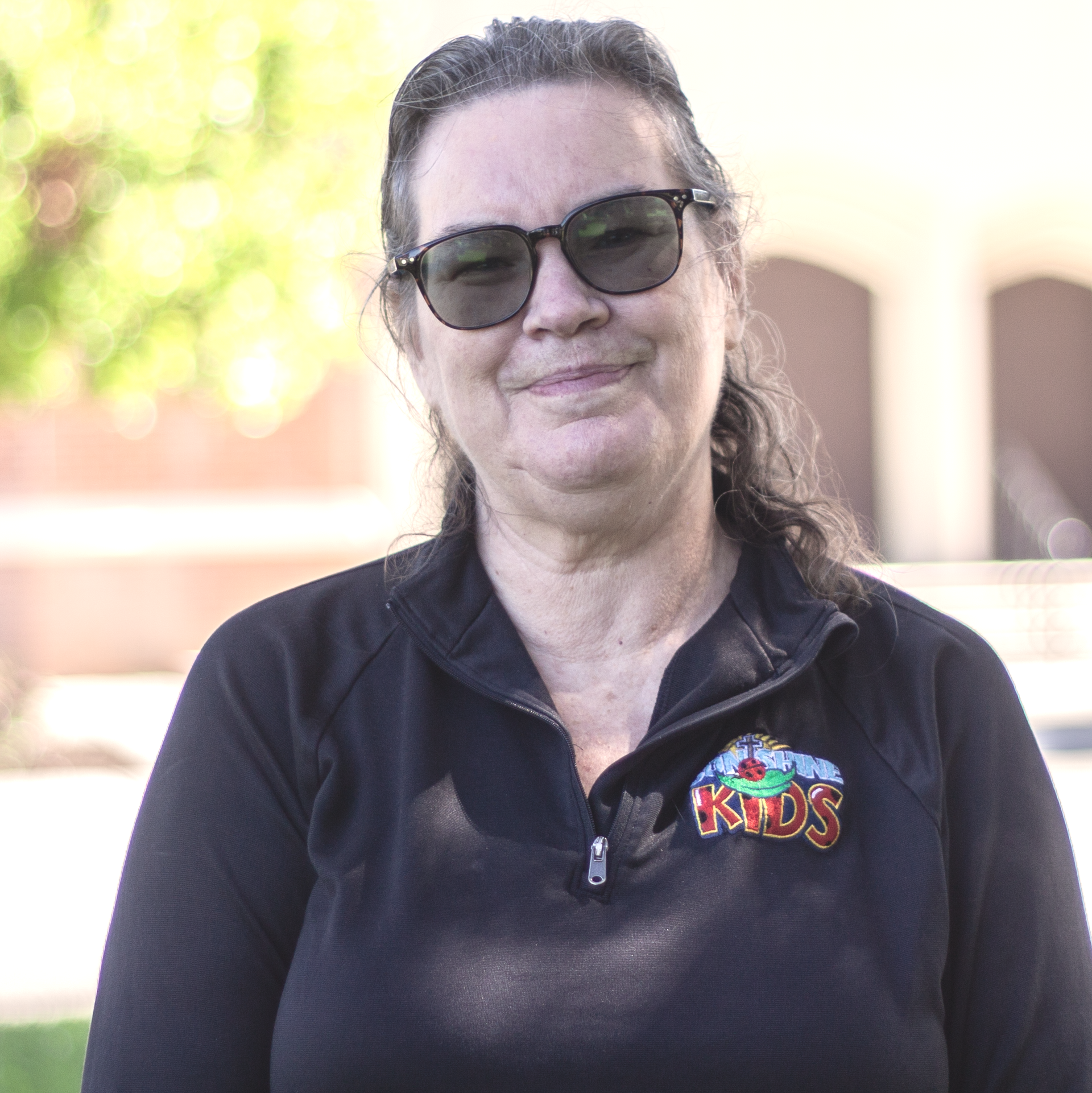 A woman in a black top with sunglasses smiling outdoors near a building.