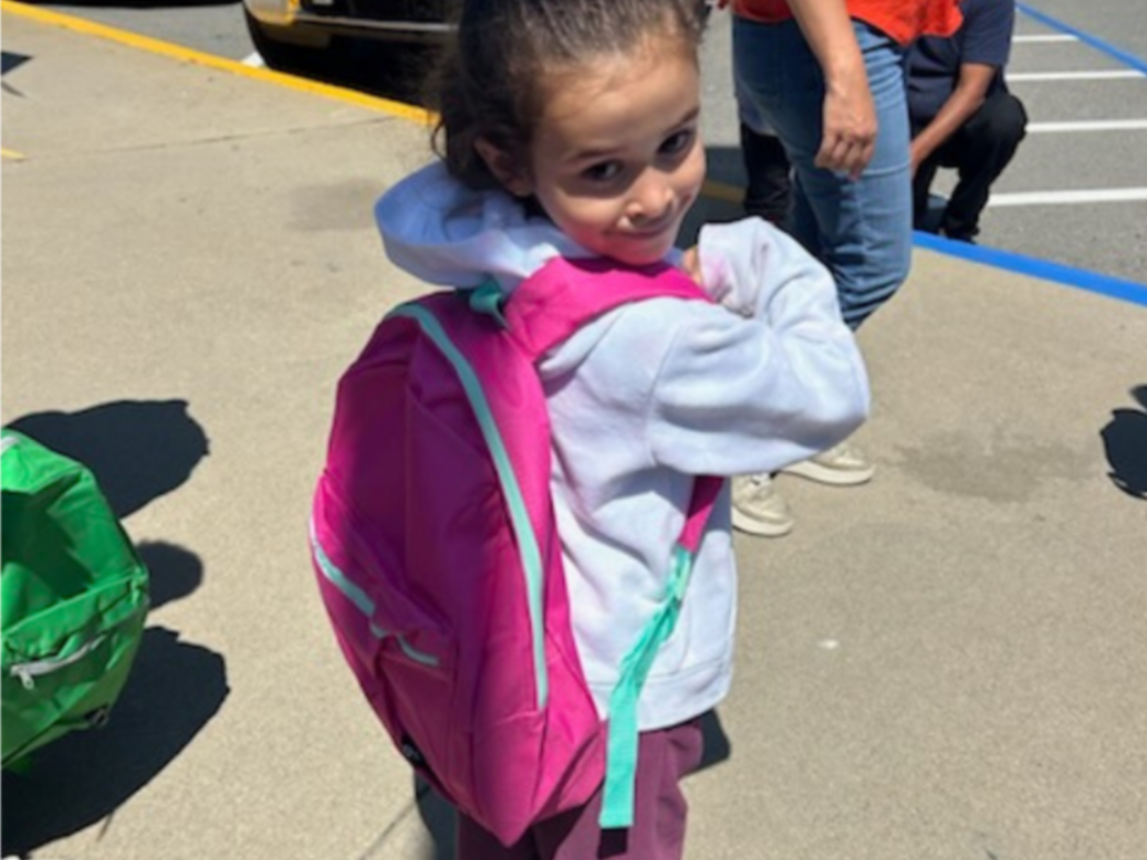 A child with a pink backpack smiles on a school playground.