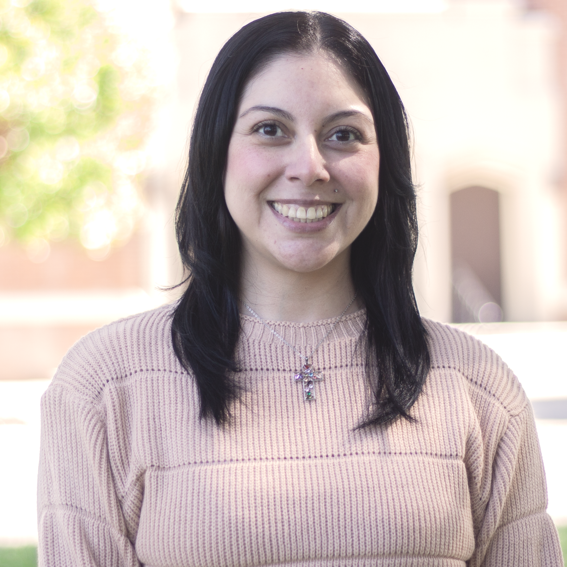 A woman with dark hair smiles in front of a building. She wears a pink sweater and a silver necklace.