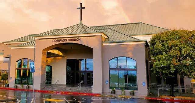 Compass Church building with a cross on top under a cloudy sky with a rainbow.