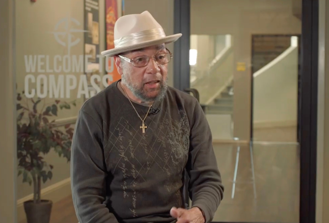 Man wearing hat and glasses speaks inside a building with