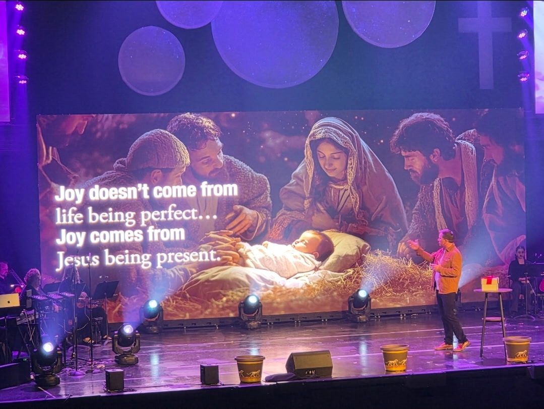 A stage with a screen displaying a nativity scene, a person in an orange shirt speaks. Text: Joy comes from Jesus.