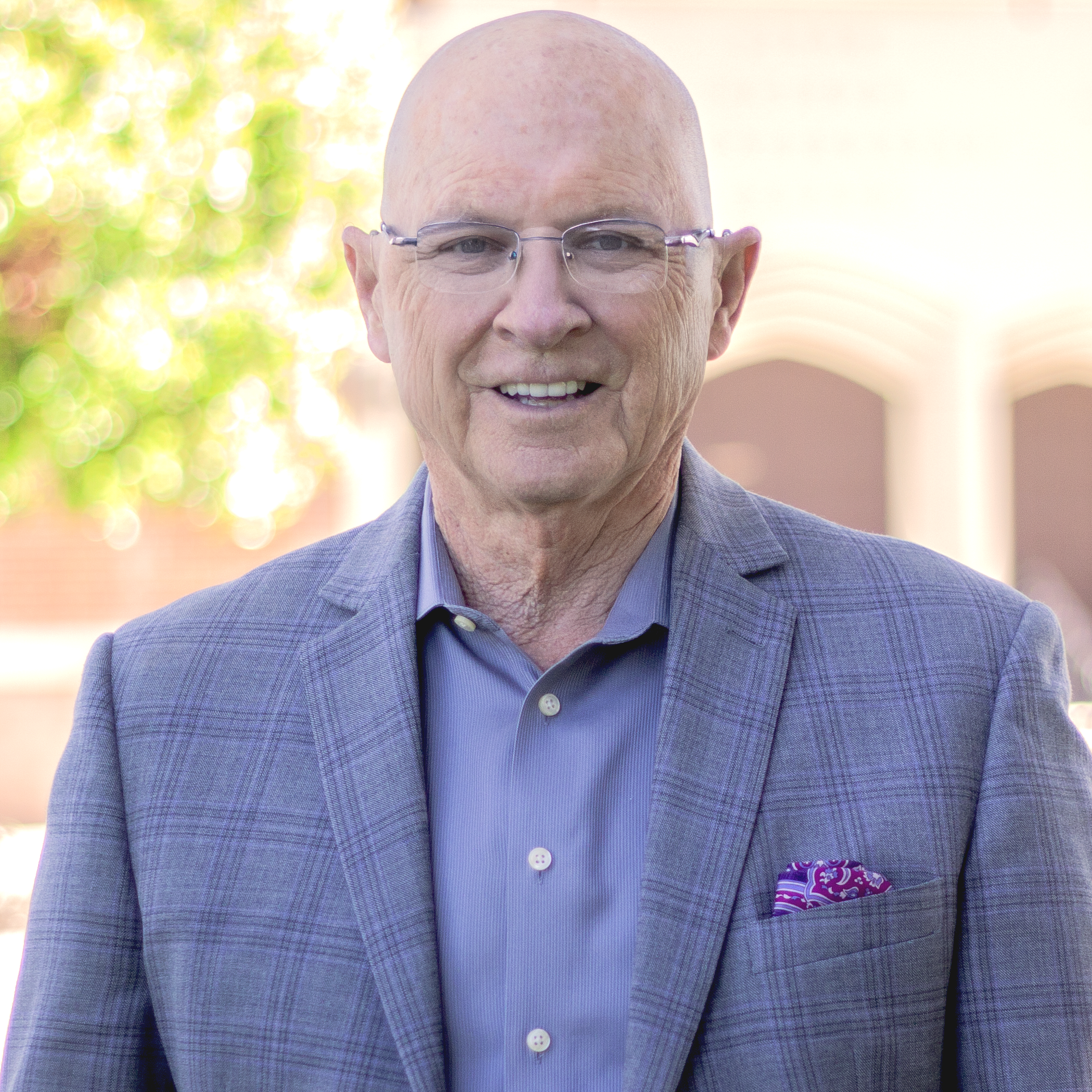 Bald man wearing glasses, blue shirt, gray blazer, and purple pocket square, smiling outdoors.