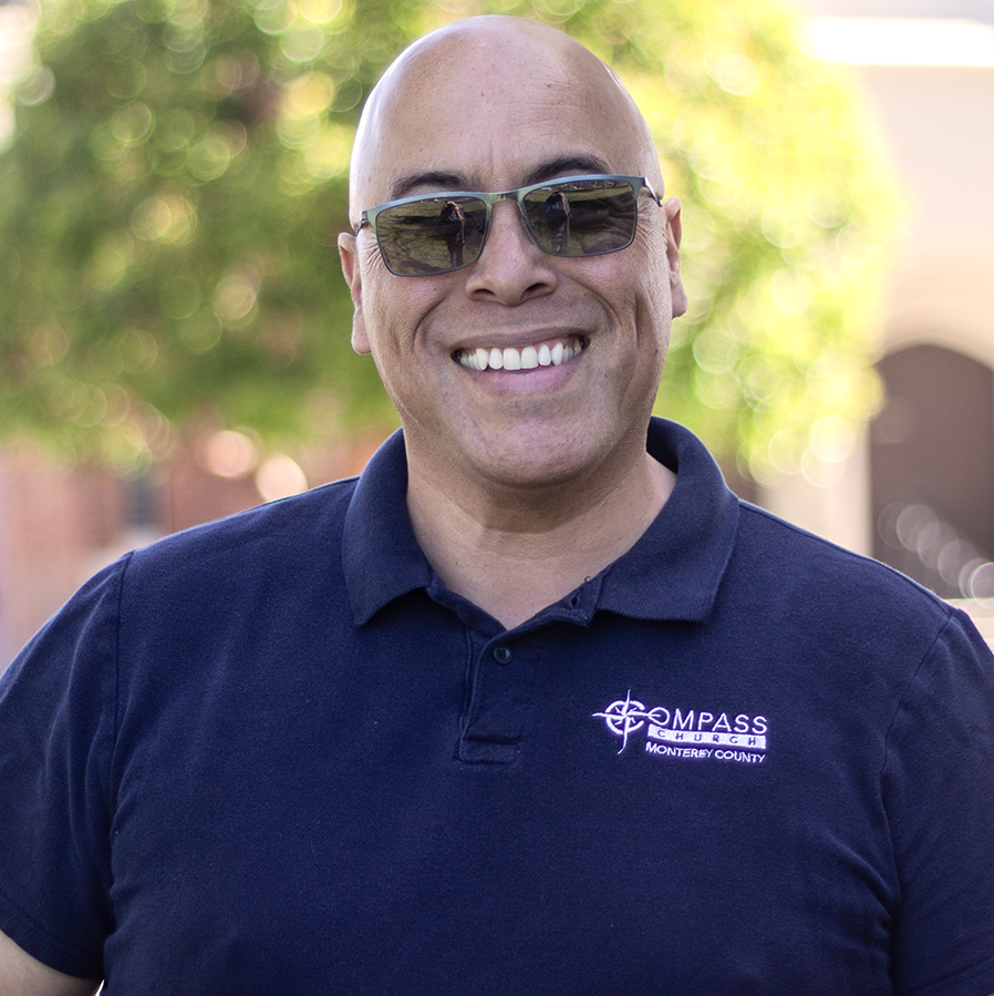 A man wearing sunglasses smiles, outdoors. Navy polo shirt with 