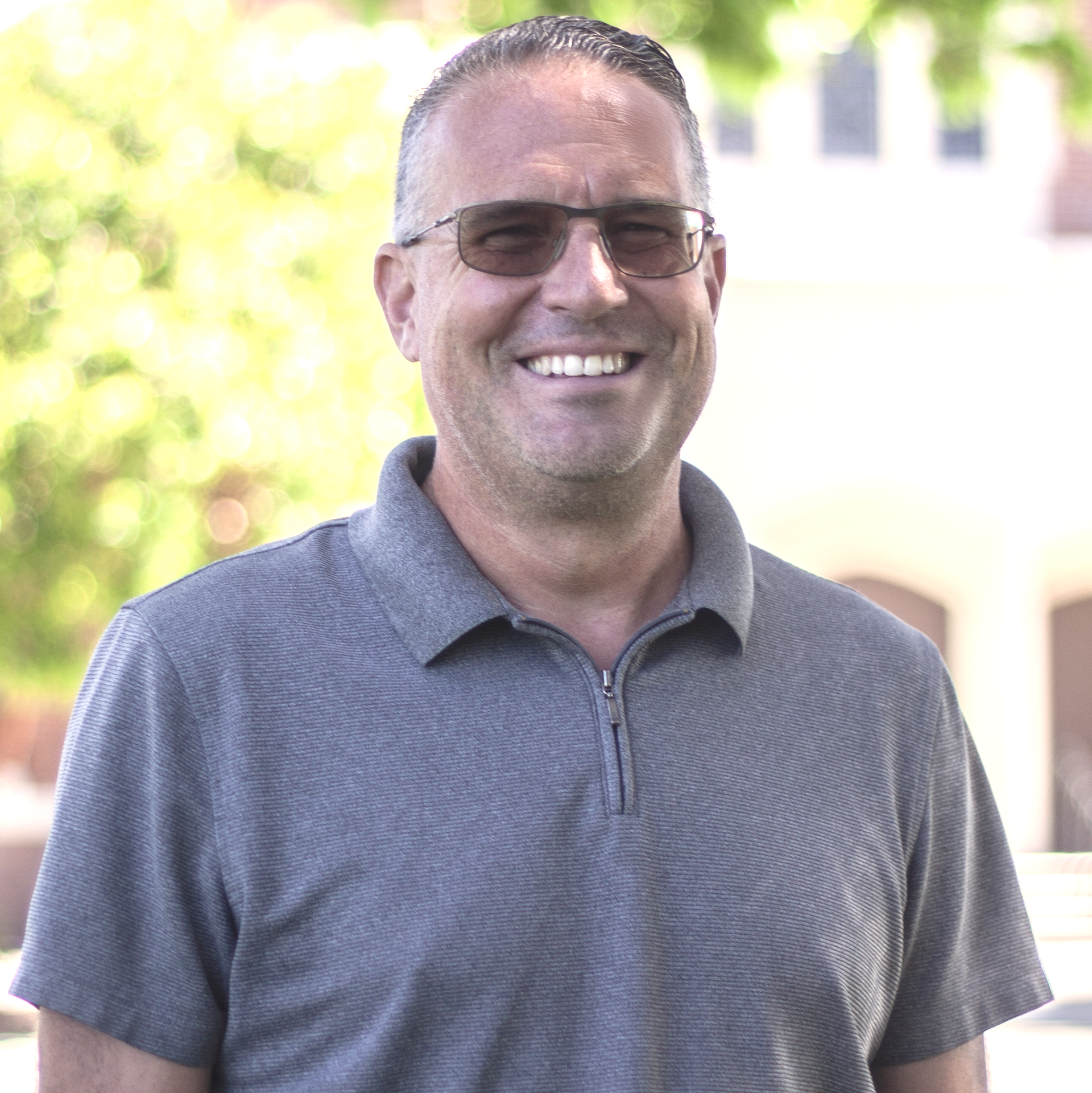 A man in a gray polo shirt and sunglasses smiles outdoors, blurred background.
