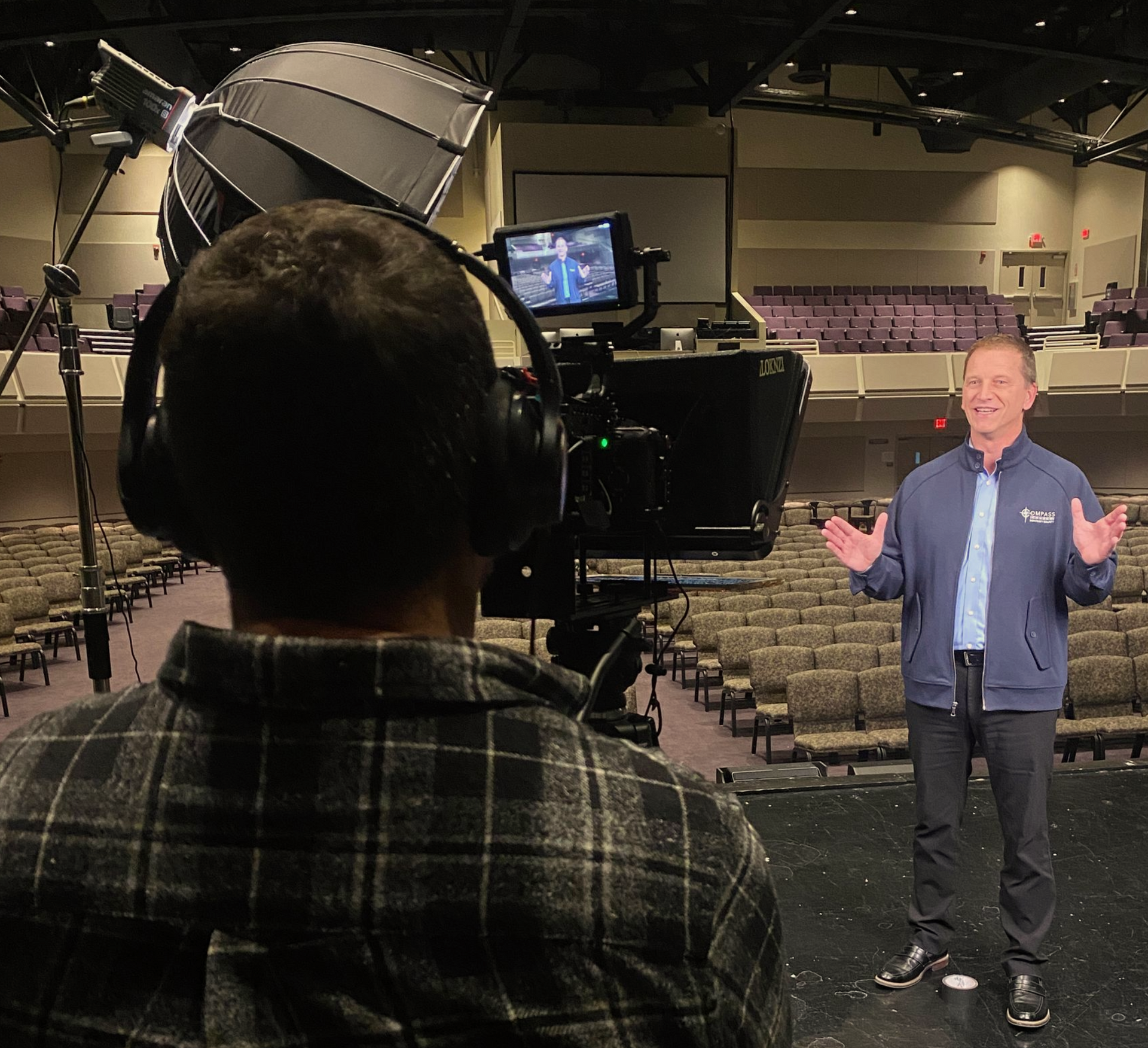 Person filming a man on a stage in an empty auditorium. The man gestures while speaking.