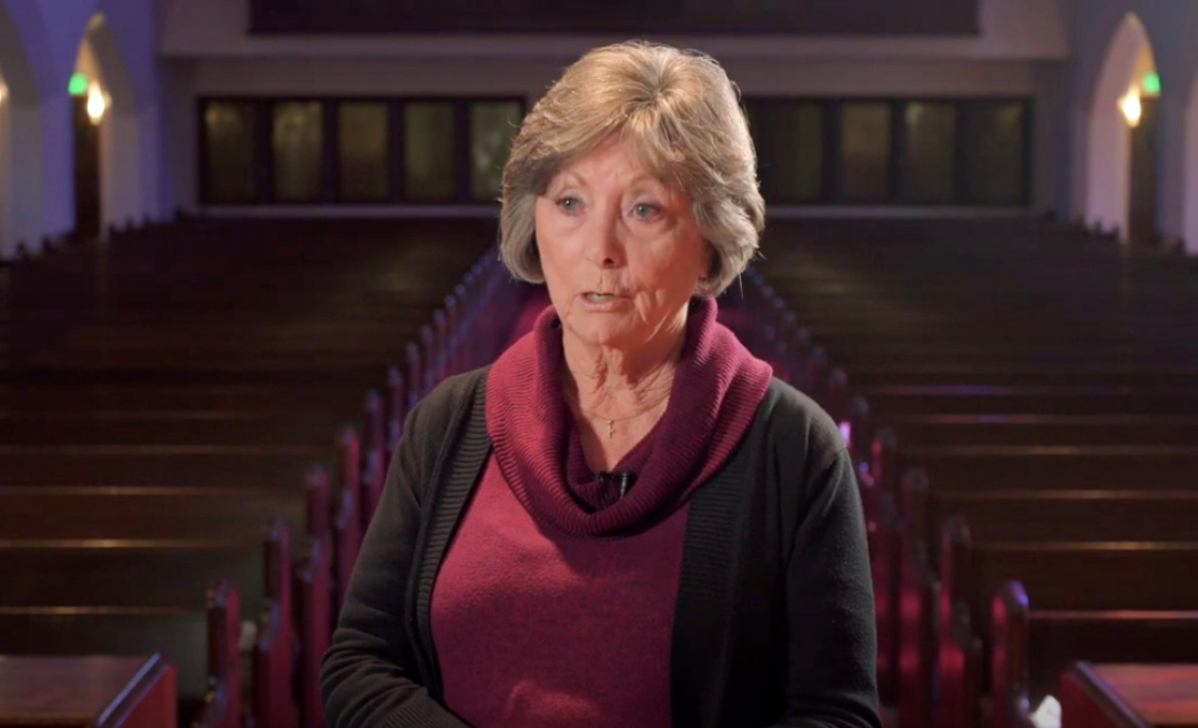 Woman in a maroon sweater stands in a church sanctuary, speaking.