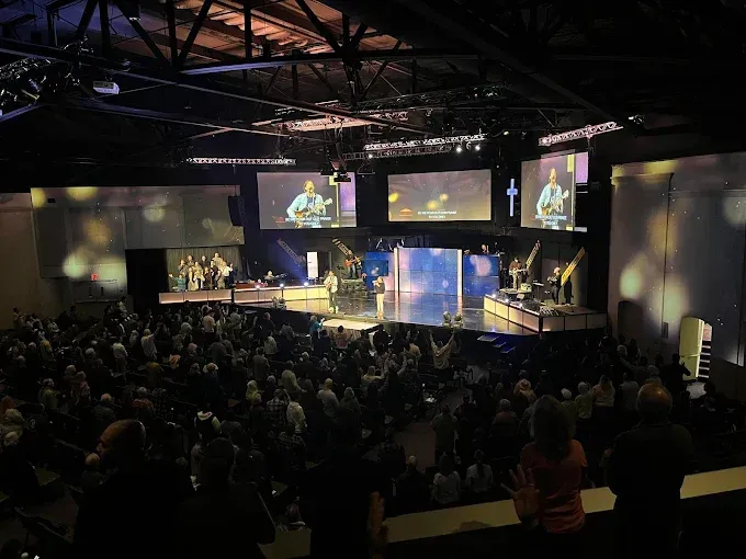 An audience at Compass Church Monterey County watches a band and a pastor on stage, with screens displaying the performance.