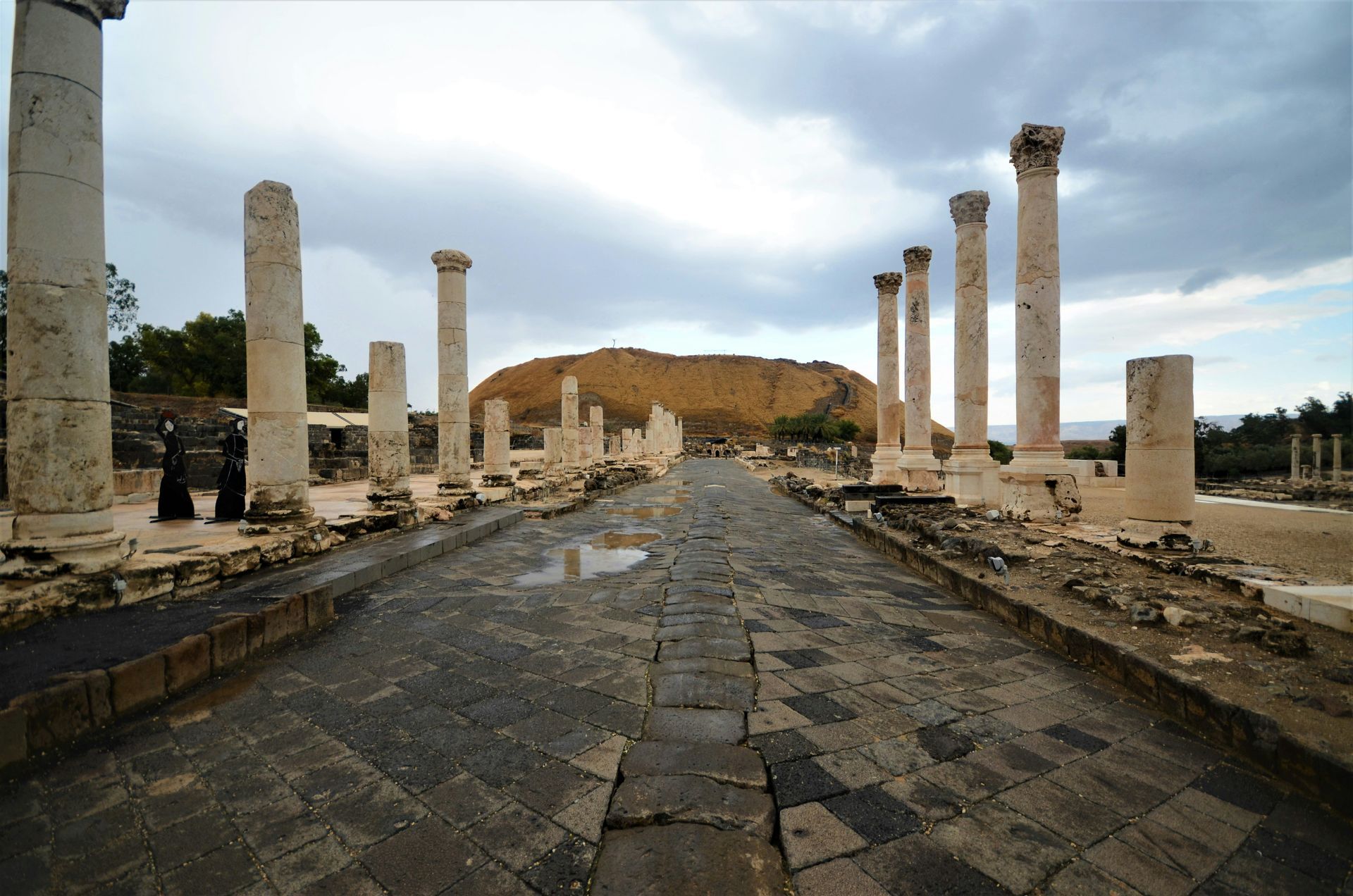 Stone road flanked by columns in ancient city ruins, with a hill in the background. Overcast sky.