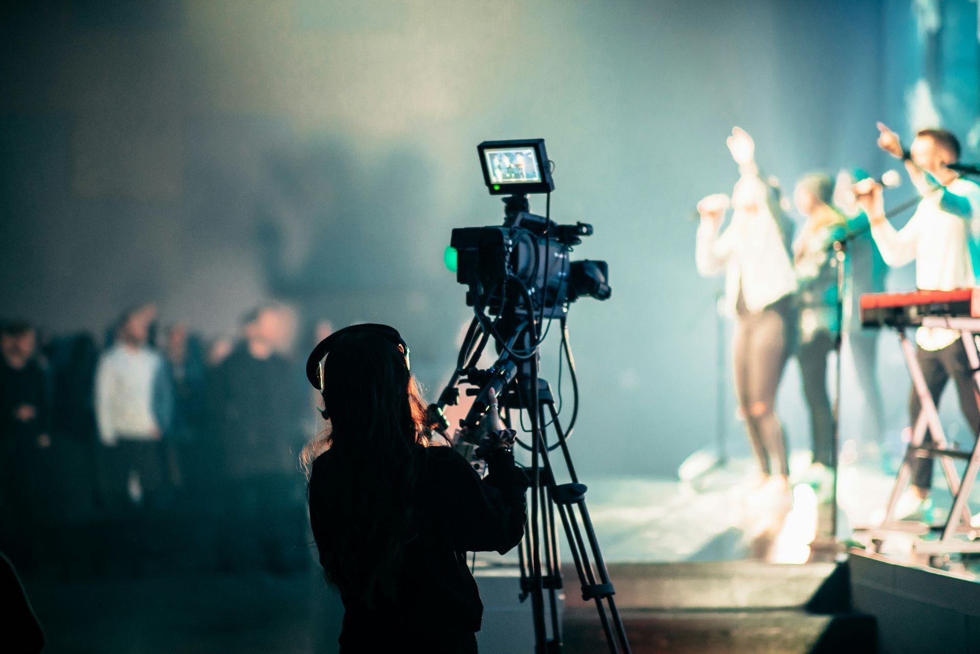 Camera operator filming a band on stage, with audience in the background and a stage lit with blue hues.