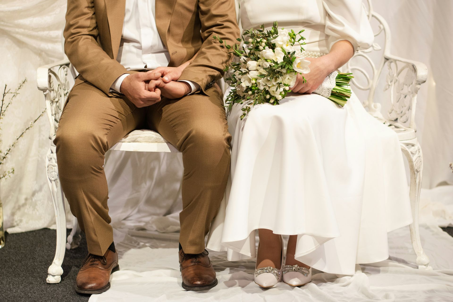 Couple in wedding attire seated on a white bench, woman holding bouquet, indoor setting.