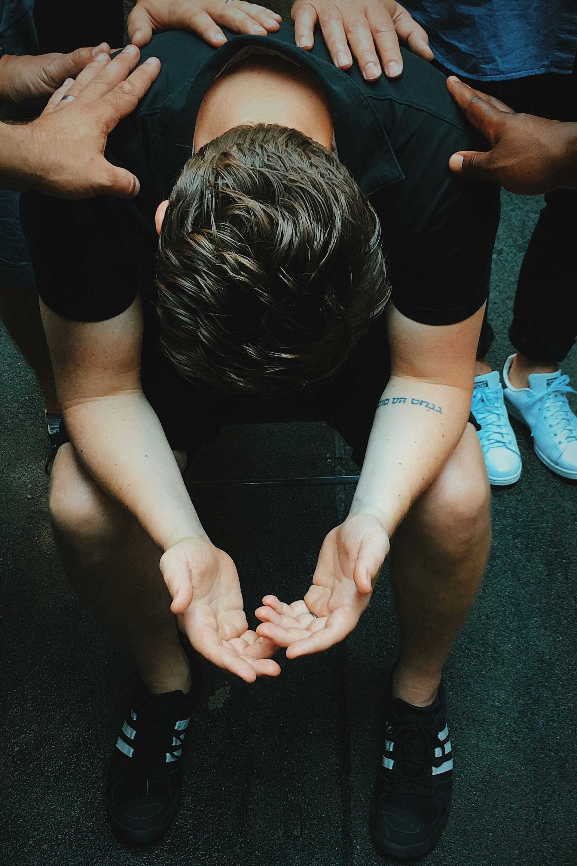 Man with head bowed, surrounded by hands. Kneeling, wearing black. Outdoors.