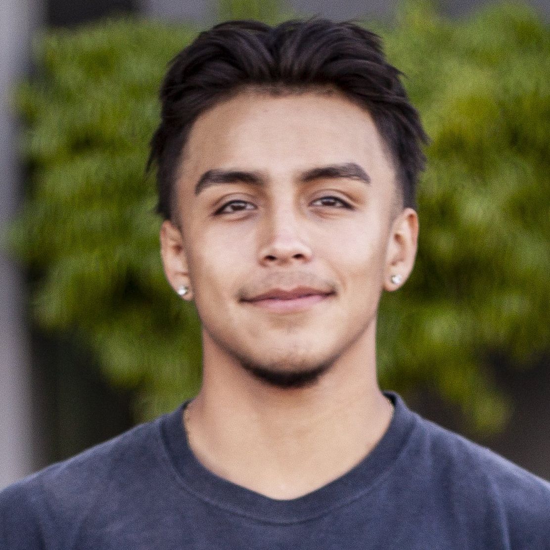 Young man with dark hair smiles, wearing a dark blue shirt and small earrings.