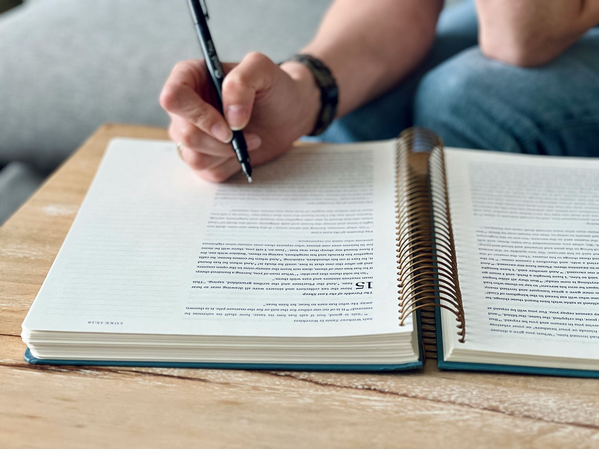 Person writing with a pen in a large open book on a wooden surface; blue jeans and a sofa in the background.