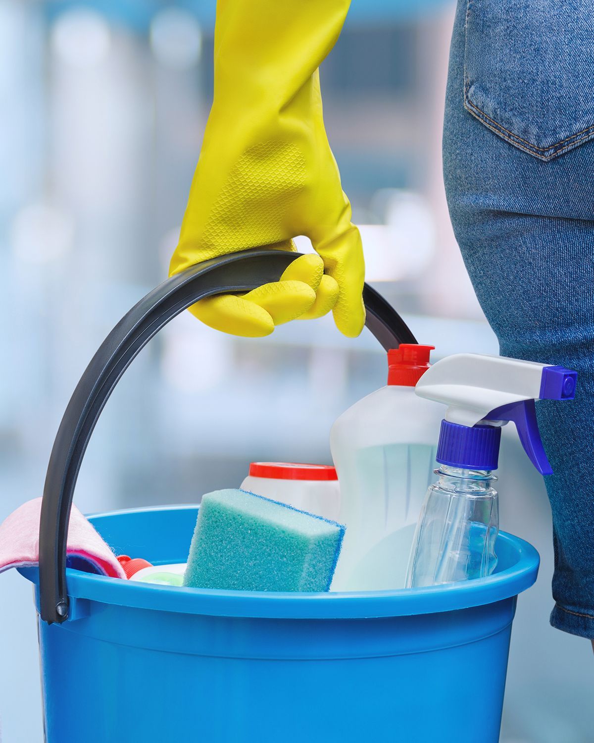 A person wearing yellow gloves is holding a blue bucket filled with cleaning supplies.