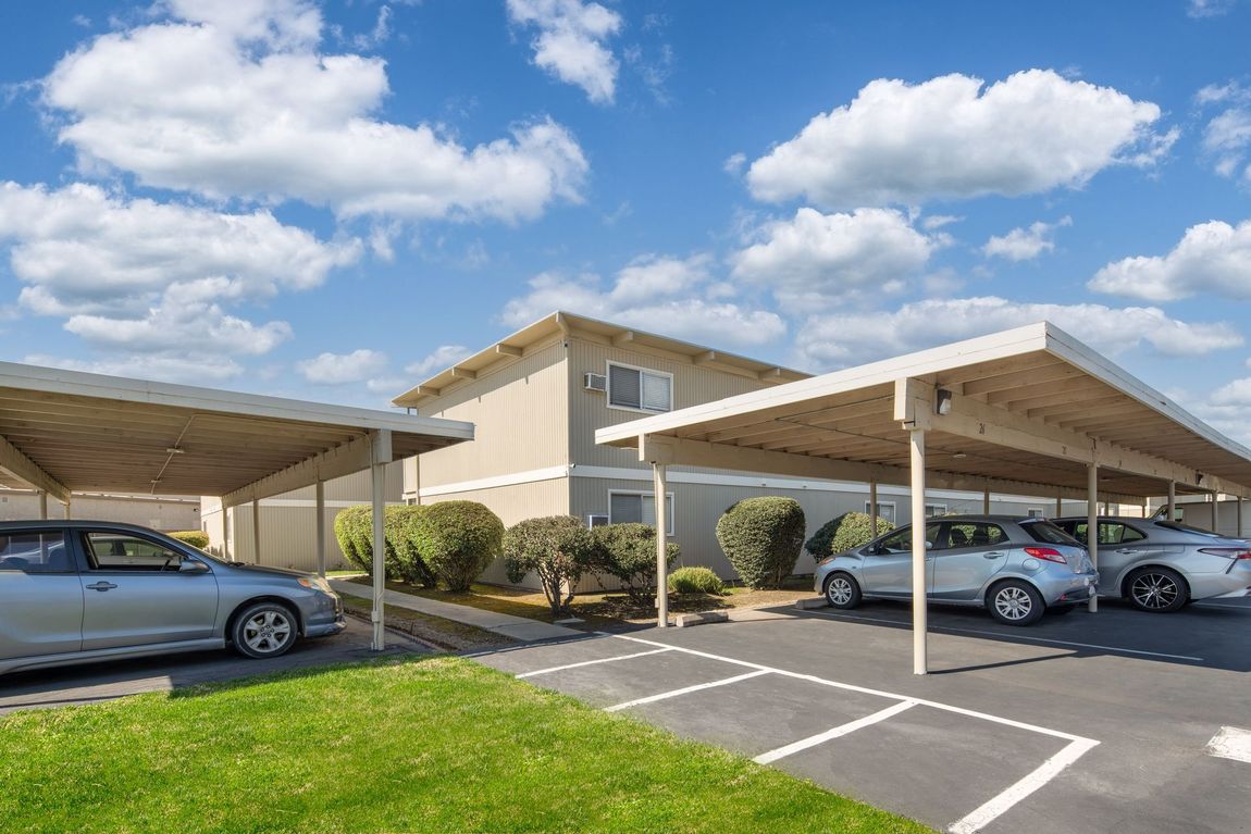 Apartment building with covered parking spaces, cars parked, blue sky with clouds, green grass.