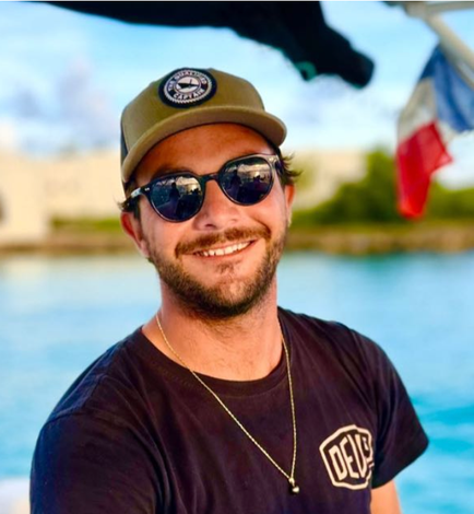 Man in sunglasses and hat smiles on a boat, blue water and a flag in the background.