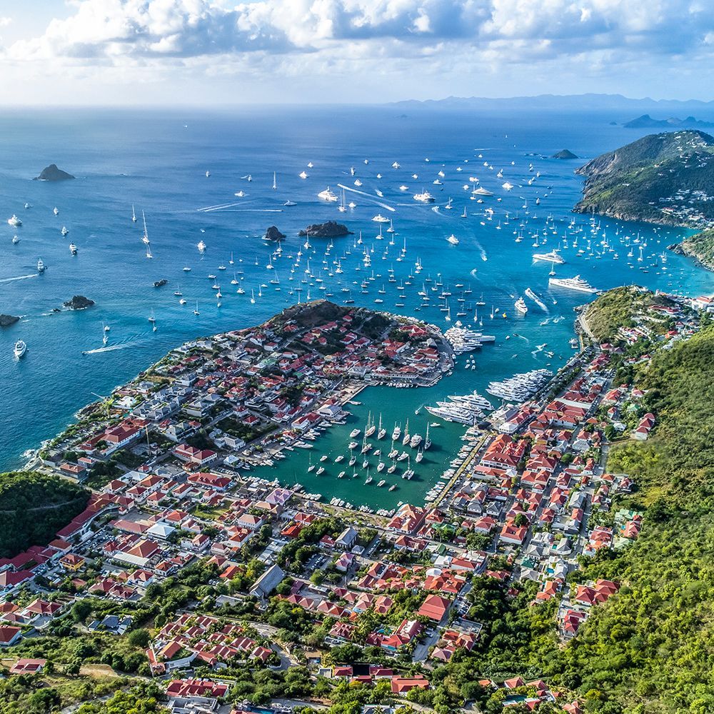 Aerial view of Gustavia, St. Barts, with boats in a harbor, buildings, and green hills. The sky is blue and cloudy.