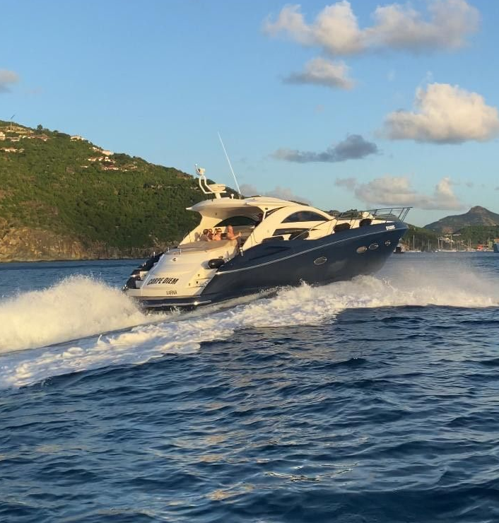 A dark blue and white yacht speeds across the water, creating a wake. Lush green hills in the background.